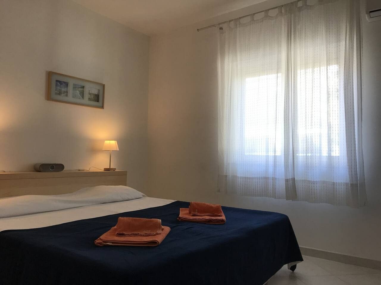 Ein Hotelzimmer mit Bett, Nachttisch, Lampe und einem Fenster mit leichtem Vorhang.
