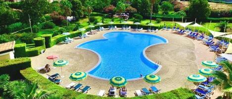A resort with a large central swimming pool, sun loungers, and palm trees.