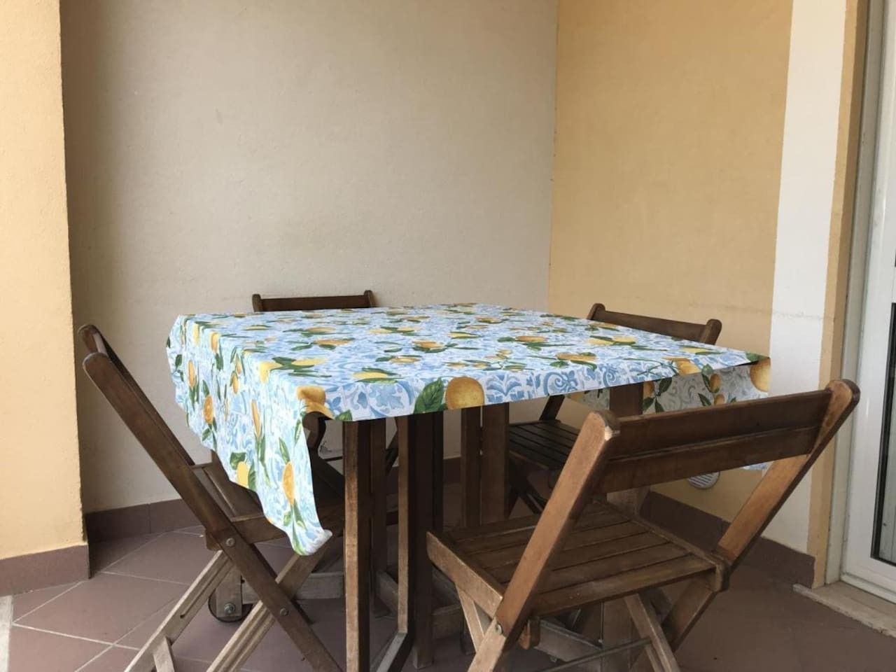 Ein Tisch mit gemustertem Tischläufer, umgeben von Holzkästen, steht auf einem Balkon mit gefliestem Boden.