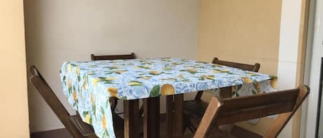 A table with a patterned tablecloth, surrounded by wooden chairs, positioned on a balcony with a tiled floor.