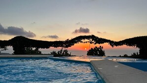 Ein Swimmingpool mit Blick auf einen Sonnenuntergang, umgeben von Bäumen und wolkenlosem Himmel.