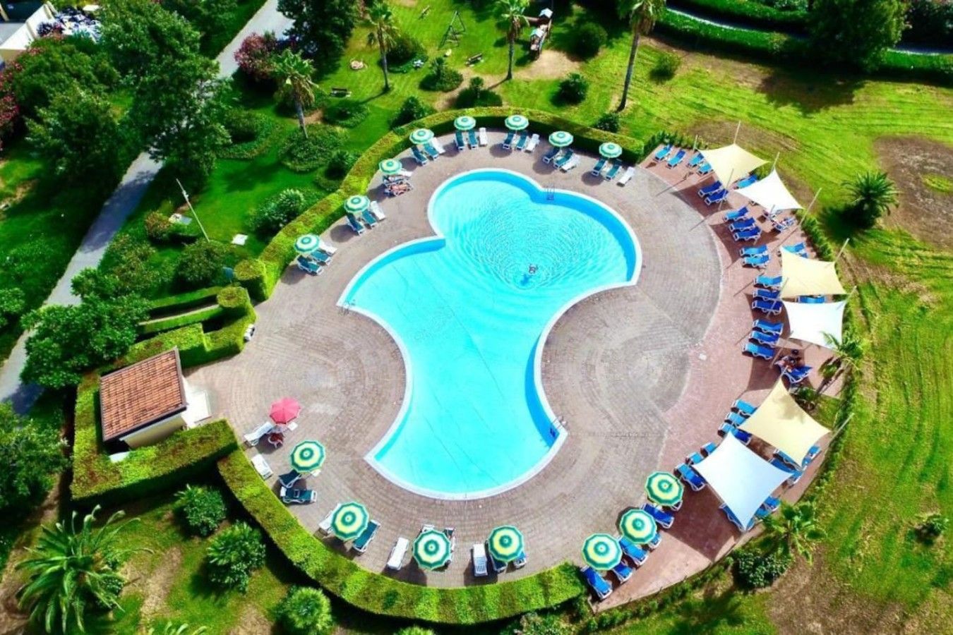 Vue aérienne d’un complexe hôtelier doté d’une piscine centrale, entourée de chaises longues et de parasols, ainsi que de jardins aménagés.