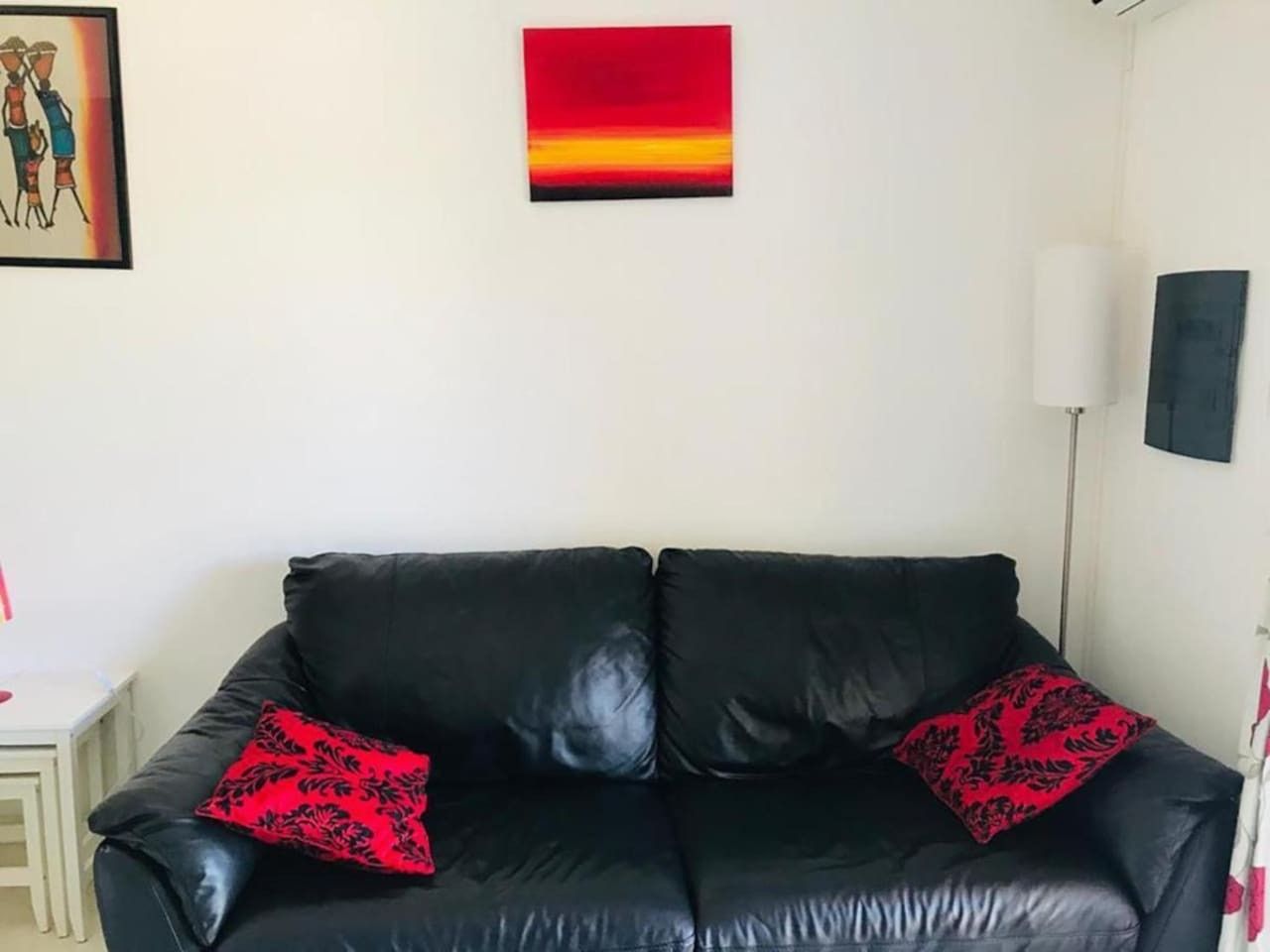 Un canapé en cuir noir agrémenté de coussins à motifs rouges, dans une pièce dont le mur est blanc et au-dessus duquel est accrochée une peinture abstraite aux couleurs rouge et jaune.