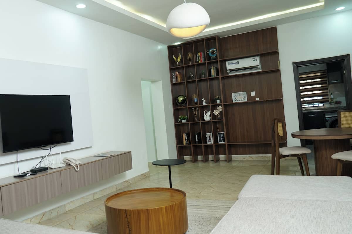 A modern living room with a flat-screen TV, a wooden coffee table, a bookshelf, and a dining area with chairs.