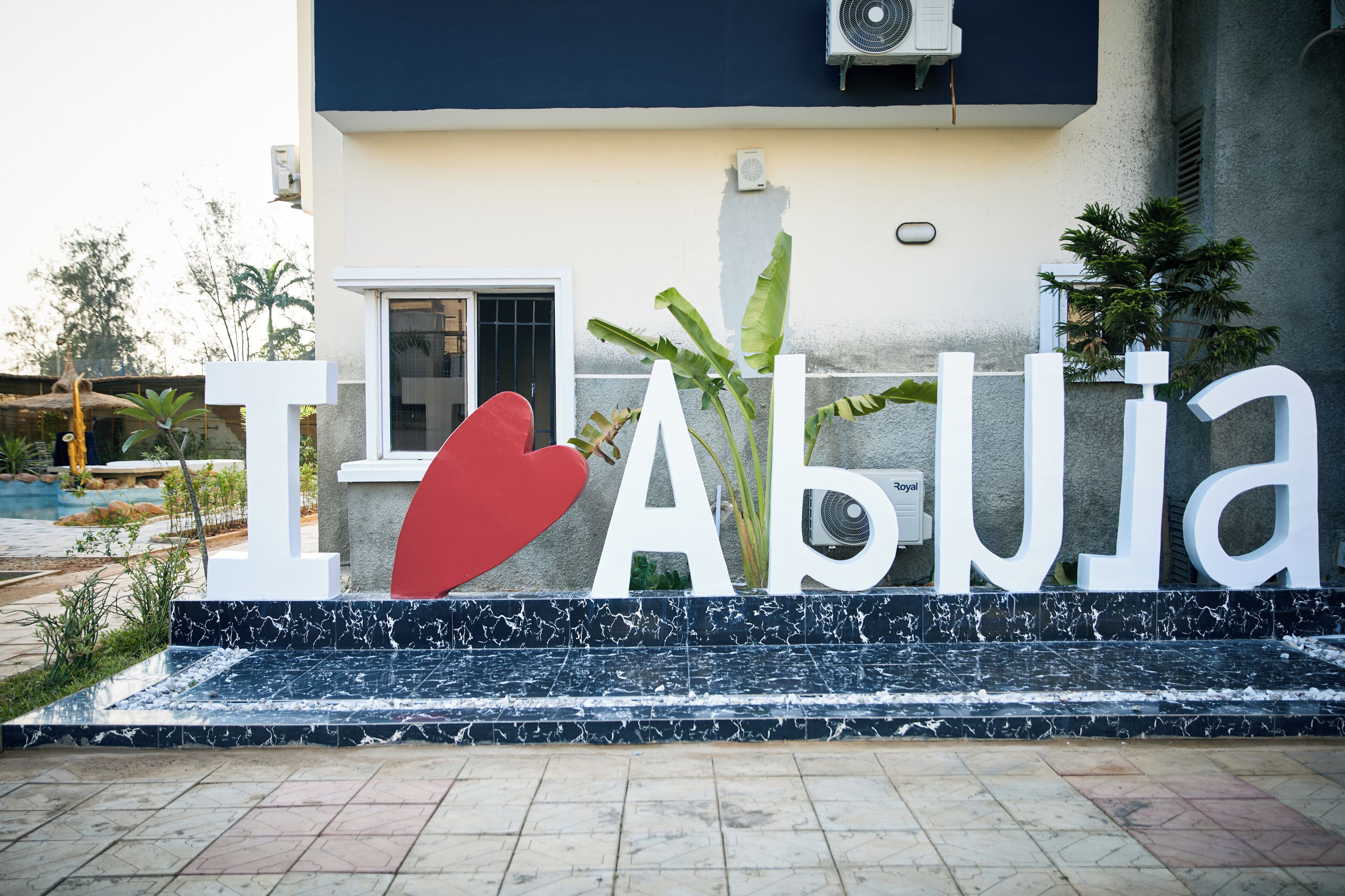 Ein Schild mit dem Schriftzug „I ♥ Abuja“ in großen weißen Buchstaben vor einer Gebäudefassade mit dunkelblauer und beiger Farbgebung.
