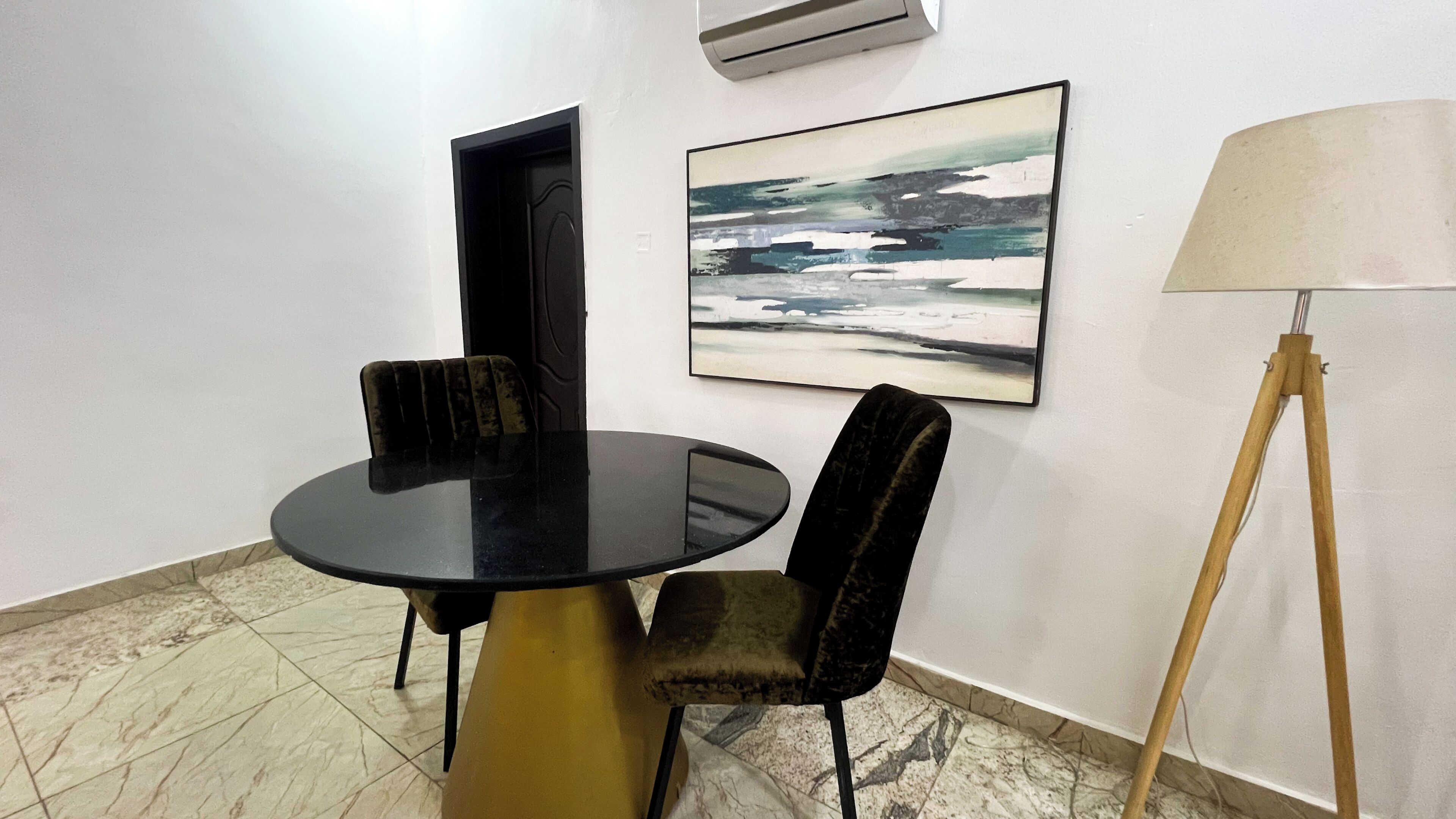 A conference room with a round table, four chairs, a standing lamp, and a painting on the wall.