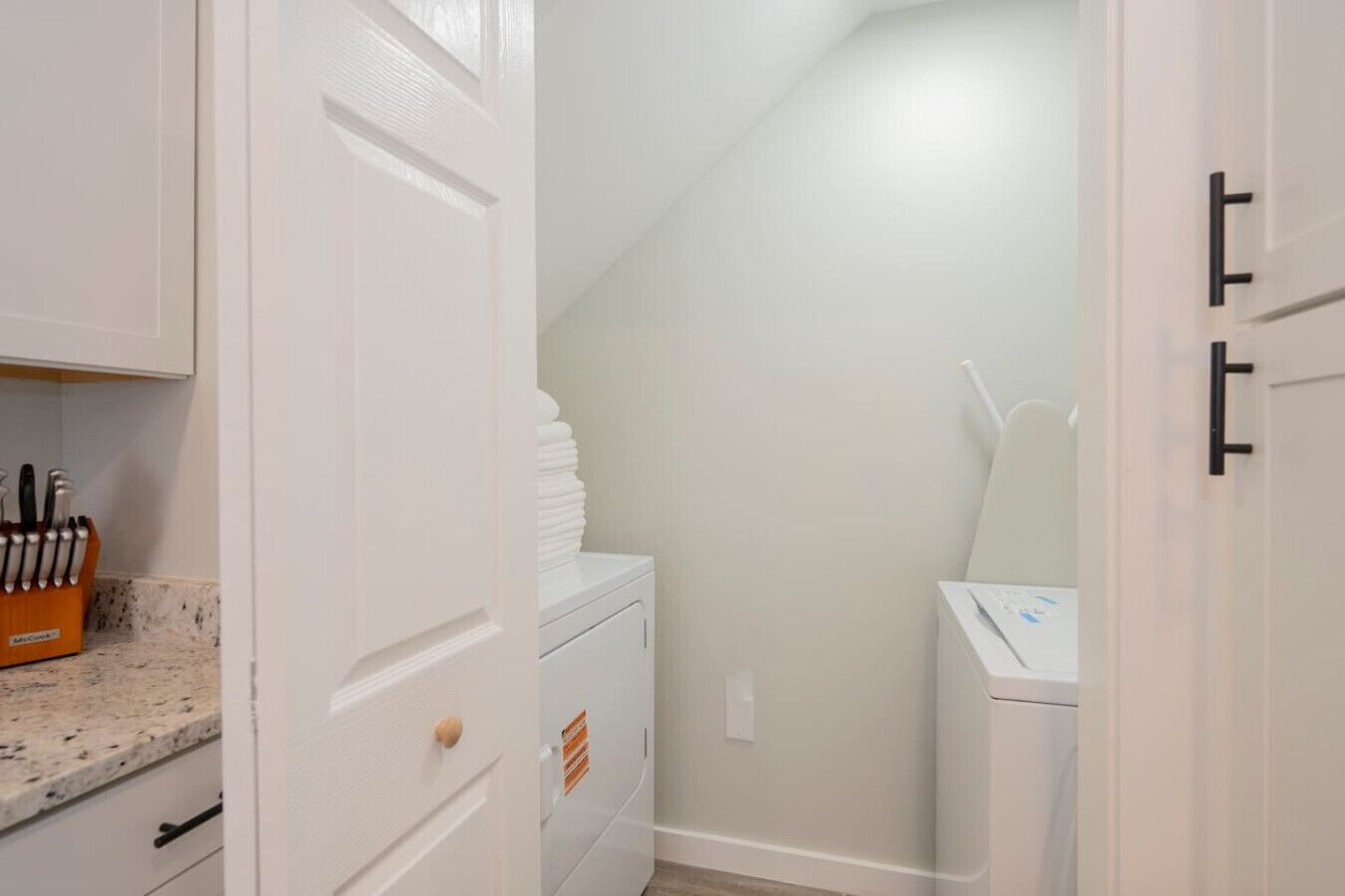 A laundry room with a stacked washer and dryer, a countertop with a knife block, and a door with black handles.