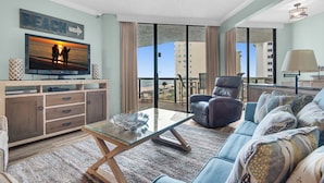 A modern living room with a large TV, a glass coffee table, a sofa, and a view of the beach through large windows.