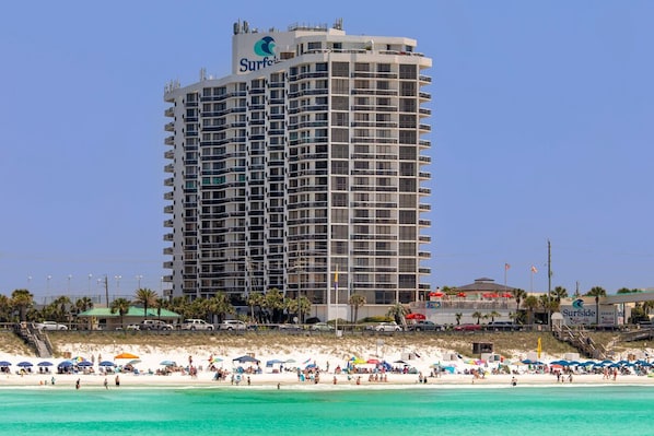 A tall hotel with a 'Surfside' sign, located near a beach with numerous umbrellas and people.