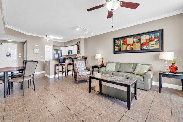 A spacious living area with a dining table, chairs, a sofa, and a coffee table. A ceiling fan and a painting are visible on the wall.