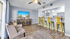A living room with a sofa, a dining area with yellow chairs, a ceiling fan, and a kitchen with white cabinets.