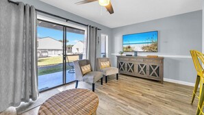 A spacious living room with a large sliding glass door leading to a patio, a ceiling fan, a flat-screen TV mounted on the wall, and a wooden cabinet.