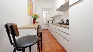 Cocina pequeña con armarios blancos, una silla de madera, un taburete y un florero con flores, además de una ventana con vistas.
