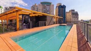 An outdoor swimming pool with a wooden pergola, surrounded by a metal fence, and a cityscape in the background. - Vossa Bossa Pop Pinheiros (São Paulo)