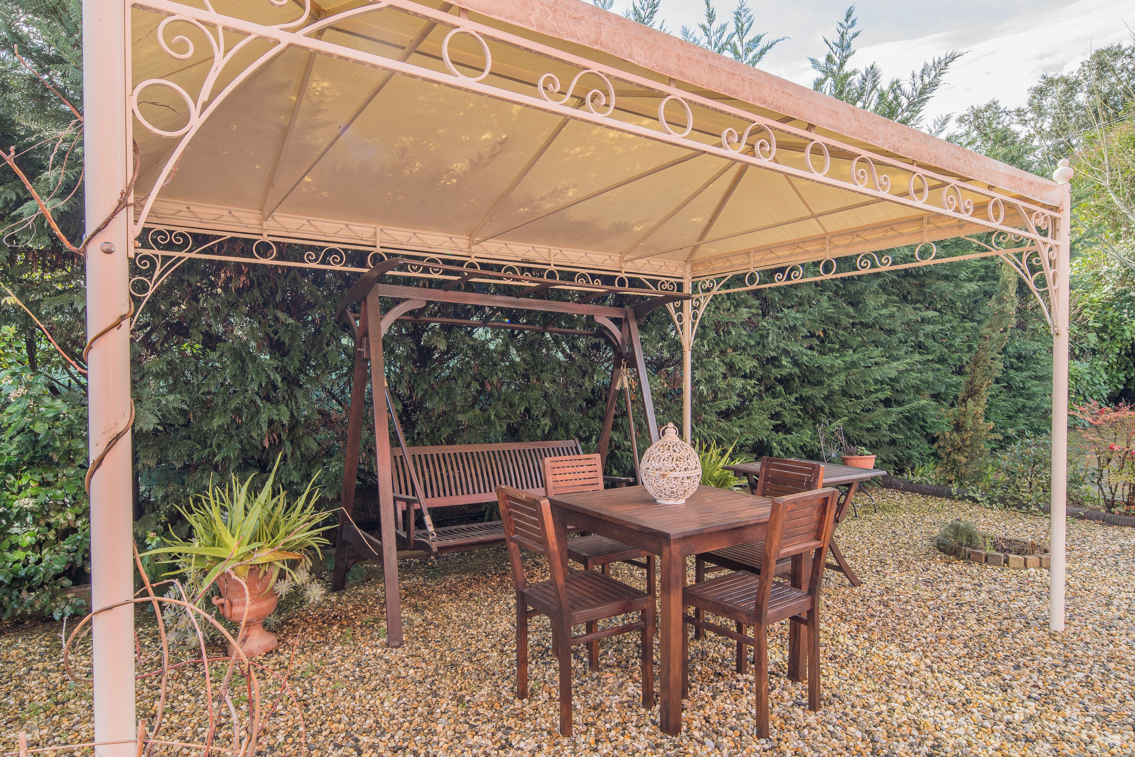 Une terrasse couverte avec une table et des chaises en bois, entourée de gravier et de plantes en pot.