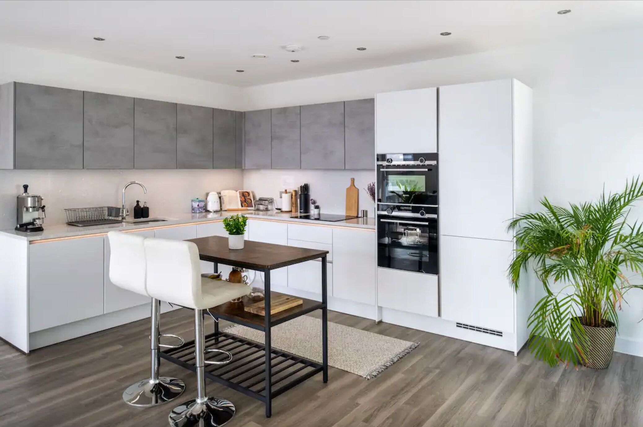 A modern kitchen with grey cabinets, a white countertop, a stainless steel sink, a built-in oven, and a bar stool.