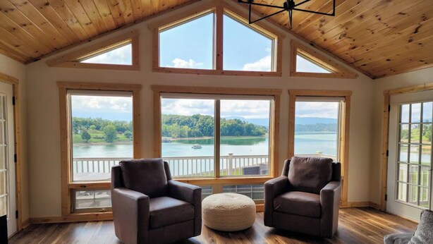 Ein geräumiges Zimmer mit großen Fenstern und Blick auf See und Wald. Es gibt zwei Ledersessel und einen runden Hocker.