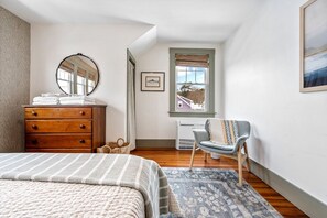 A bedroom with a bed, dresser, chair, window, and artwork.