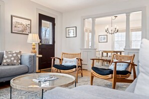 A living room with a sofa, two wicker chairs, a coffee table, and a dining table with chairs. There are framed pictures on the walls and a chandelier. - Steps away from Downtown OGT! (Ogunquit)