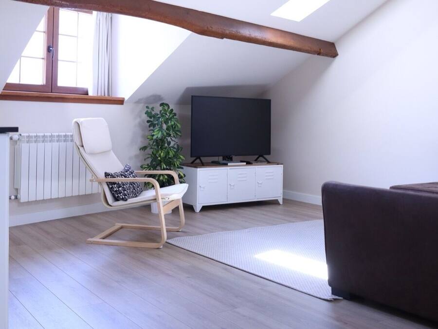 A modern loft apartment with a sloped ceiling, a large window, a flat-screen TV, a sofa, a chair, a plant, and a white cabinet.