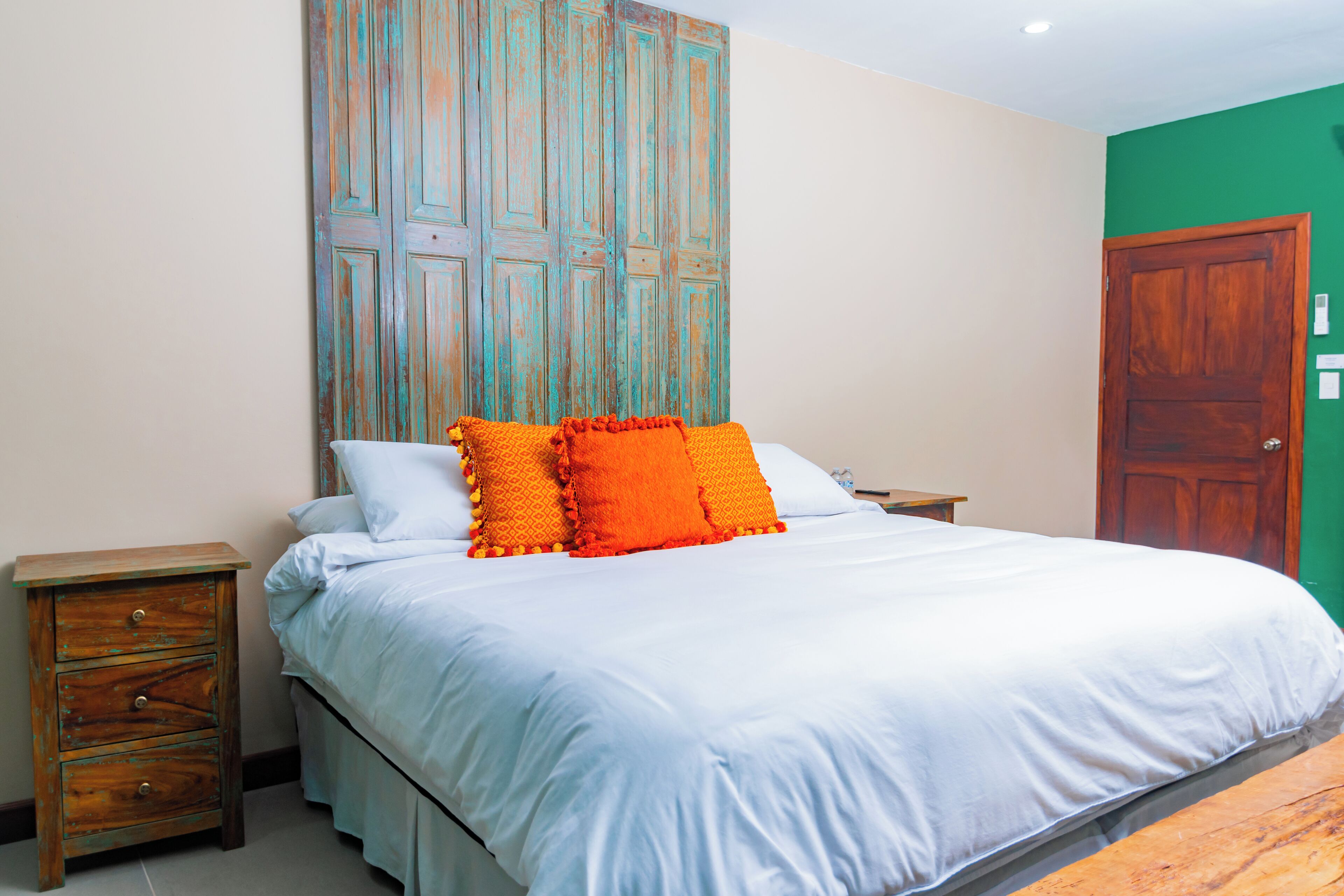 Quarto com cama, mesas de cabeceira de madeira, parede verde e porta de madeira.