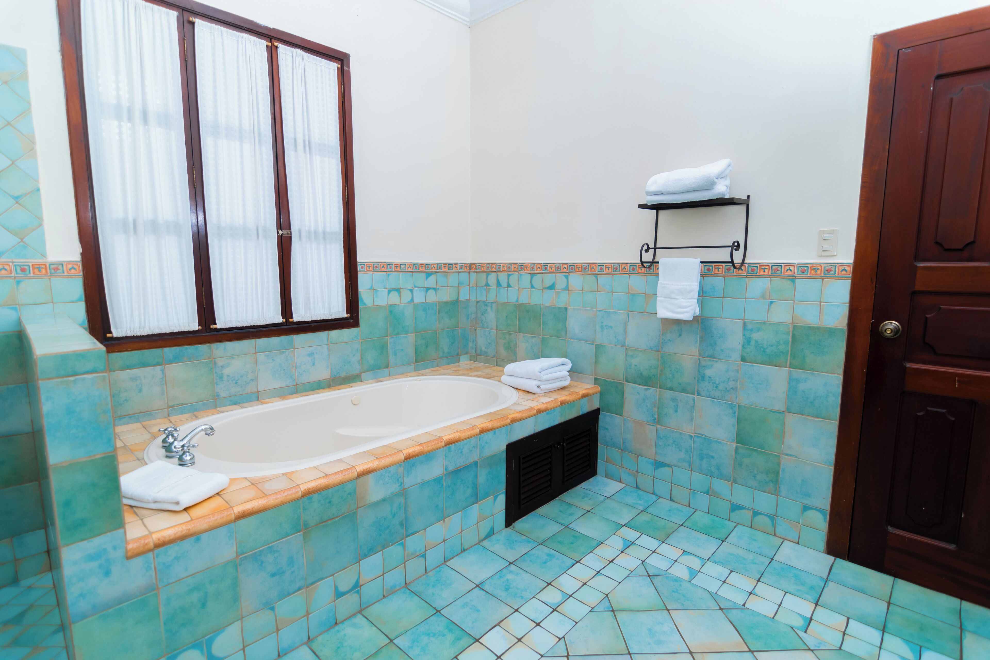 Une salle de bain avec une baignoire, des murs carrelés, une fenêtre avec des rideaux, un porte-serviettes et une porte.