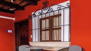 A small outdoor seating area with a round table and two chairs, a window with metal bars, and a red wall.