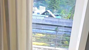 Una habitación con cama, ventana con cortinas y vista a un balcón con barandilla y vegetación.