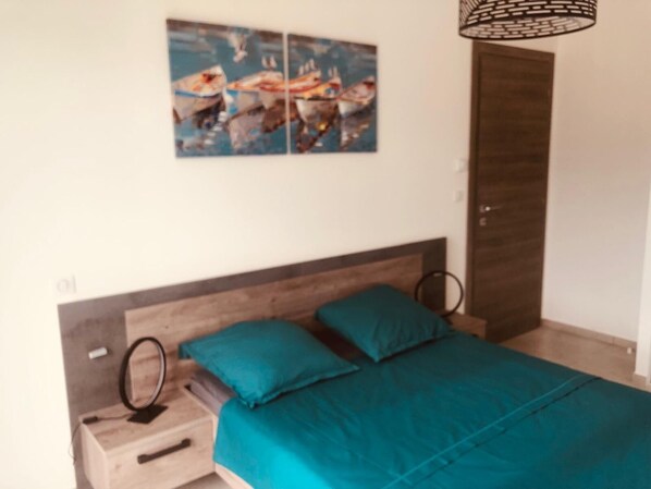 A bedroom with a wooden headboard, a bed with teal bedding, a bedside table with a ring light, and a wall-mounted artwork of boats. - Apartment T2 Île Rousse (L'Île-Rousse)