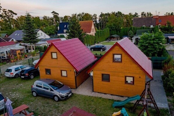A residential area with two wooden houses, each with a red roof, surrounded by greenery and parked cars.