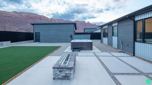 A modern outdoor patio with a stone fire feature, a hot tub, and a view of a mountain range. - Crimson Place - Moab Home with a Private Pool! (Moab)