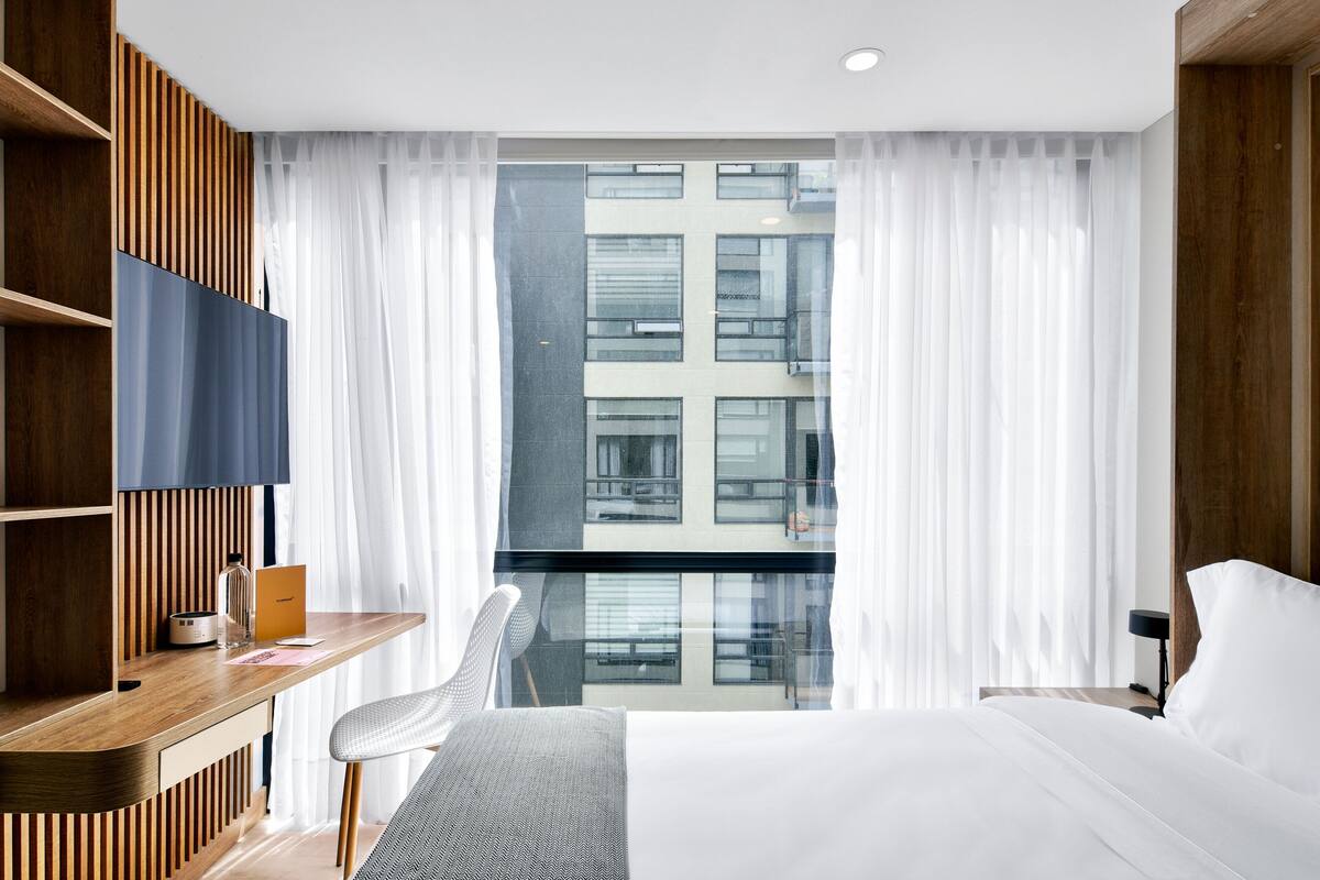 A modern hotel room with a large bed, a desk with a laptop, a chair, and a view of a building through sheer curtains.