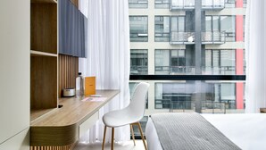 A modern hotel room with a wooden desk, a white chair, a bed with a patterned blanket, and a large window offering a view of a multi-story building. - Nuo by Wynwood House  (Bogotá)