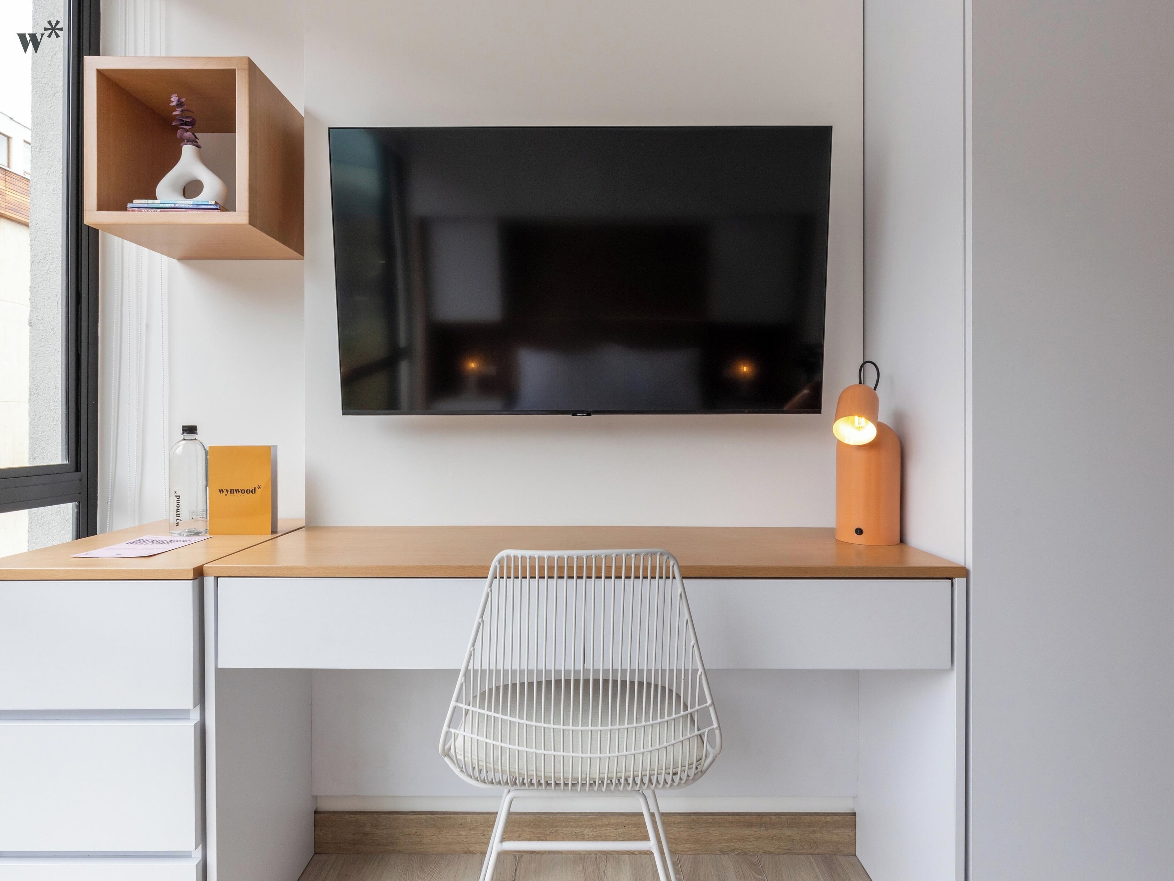 Un espace de travail moderne équipé d’un téléviseur à écran plat, d’une lampe de bureau, d’une chaise blanche et d’une étagère en bois agrémentée d’objets décoratifs.