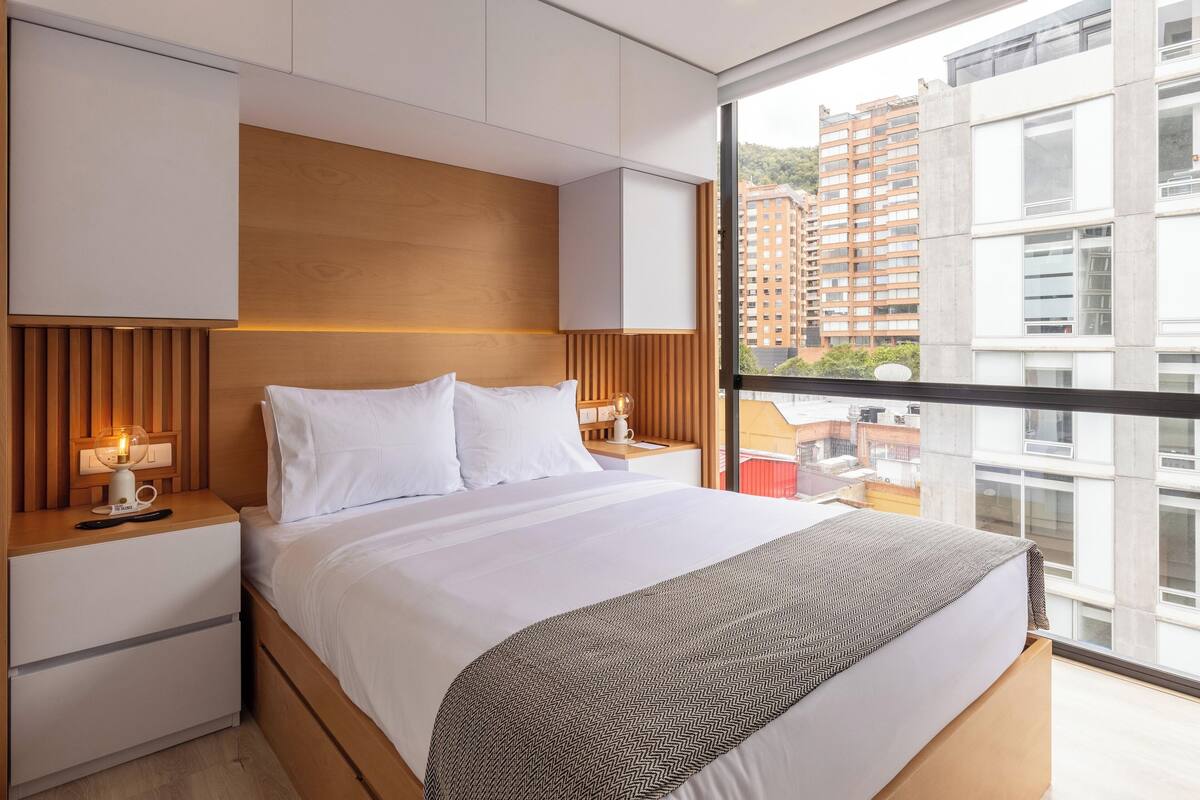 A modern bedroom with a large bed, wooden headboard, bedside tables with lamps, and a large window offering a city view.