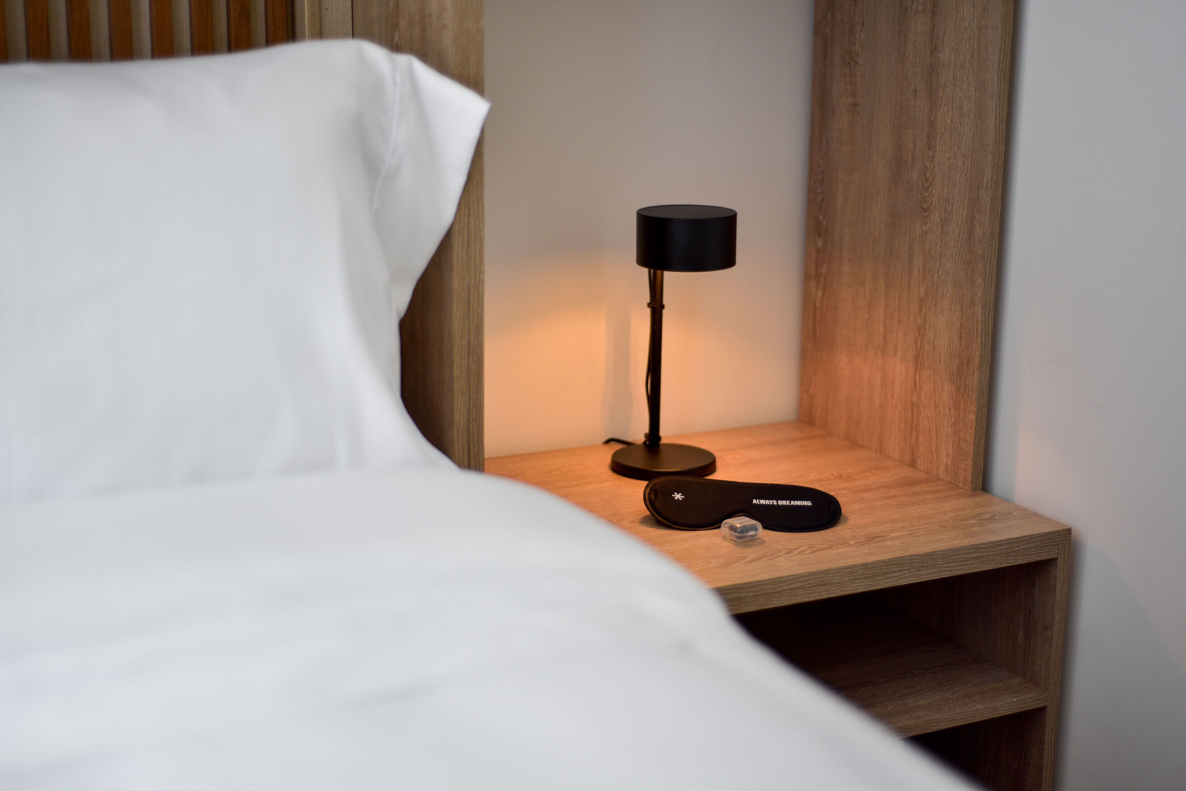 A neatly made bed with white sheets, a black desk lamp, and a small black object on the nightstand.
