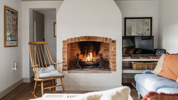 A living room with a brick fireplace, a wooden rocking chair, a blue sofa, and a television on a shelf. - A Pretty Norfolk Cottage (Field Dalling)