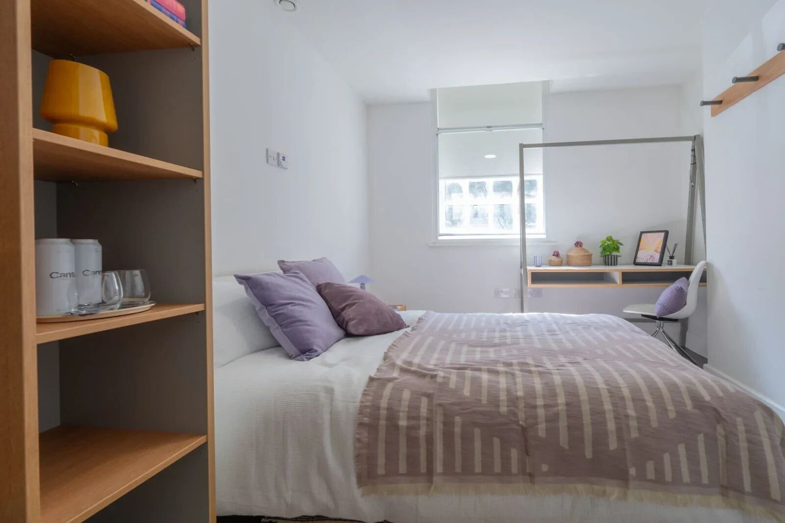 A bedroom with a bed, a desk, a chair, a window, and a shelf with items.