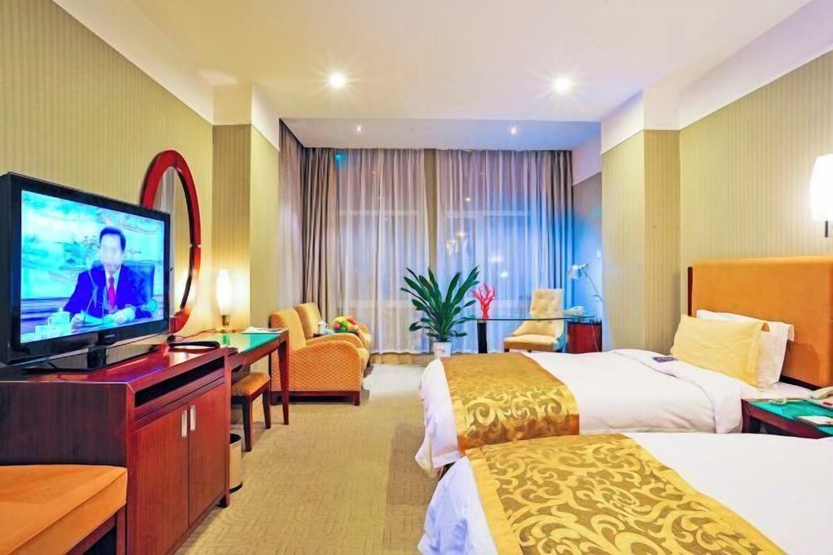A hotel room with a television, two beds, a desk, a chair, a potted plant, and a window with curtains.