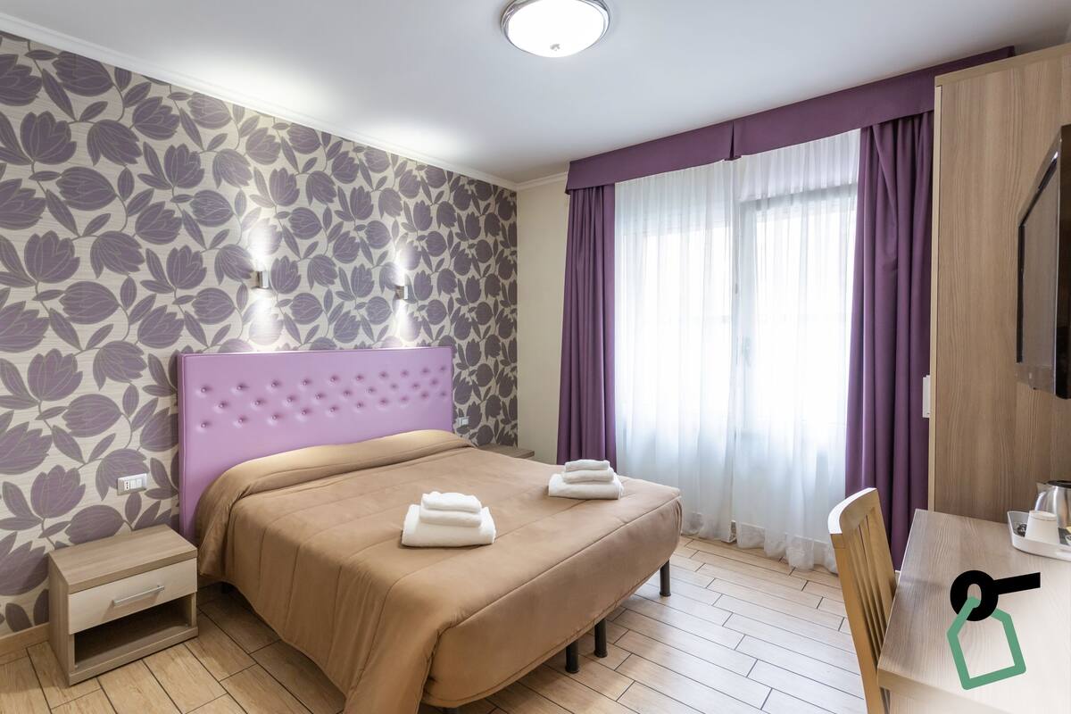 A hotel room with a bed, a nightstand, a desk with a chair, and a large window with curtains.