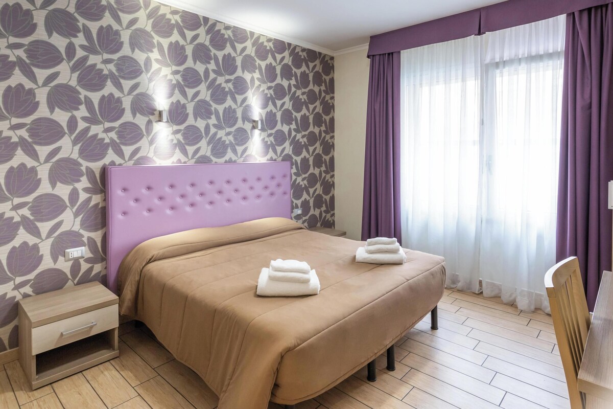 A hotel room with a bed, a nightstand, a desk with a chair, and a large window with curtains.