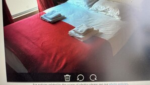A bed with a red bedspread and white pillows, a folded towel on the bed, and a window with curtains.