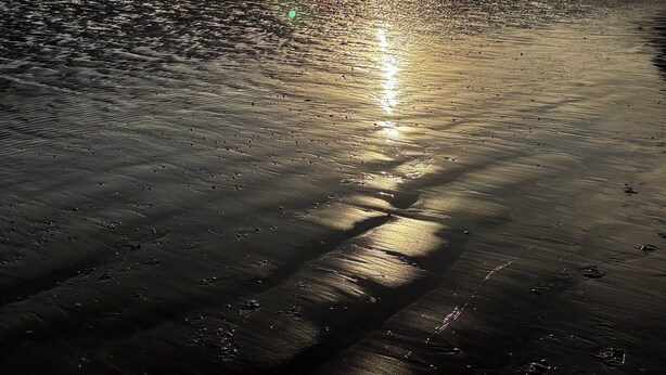 A beach with the sun reflecting on the water, a clear sky, and distant mountains.