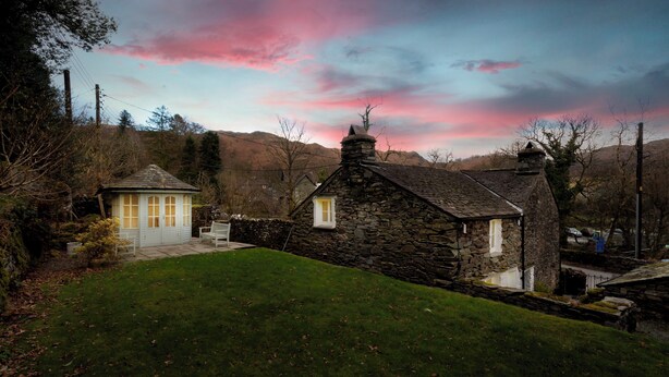 A stone cottage with a small porch and a bench, surrounded by a grassy yard and trees.