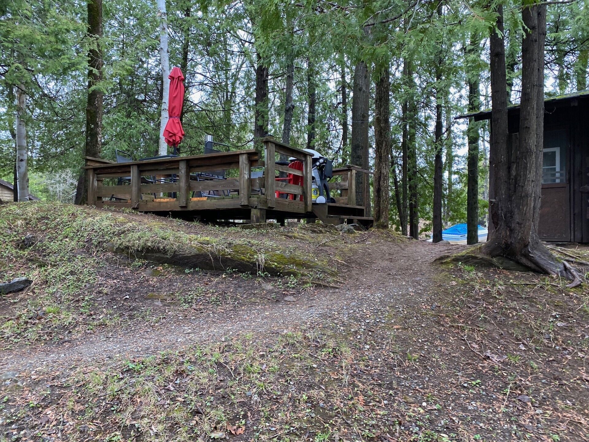Une terrasse en bois avec des sièges et un parasol rouge, entourée d’arbres.