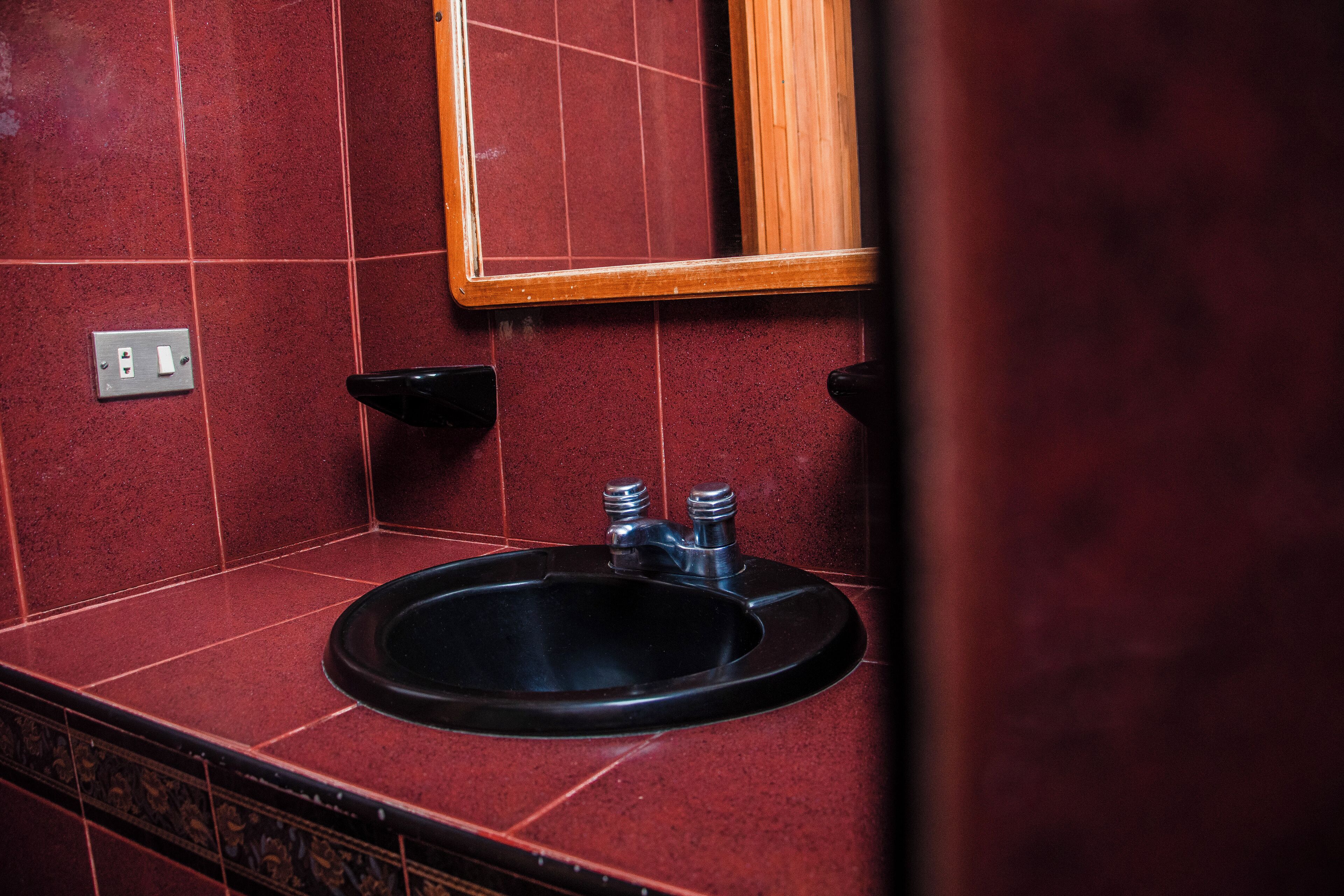 A bathroom with a black sink, red tiled walls, a wooden-framed mirror, and a wall-mounted light switch.