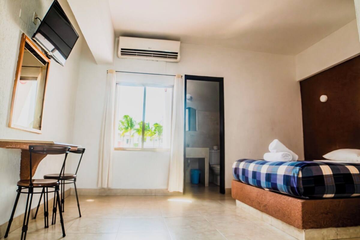 A hotel room with a bed, a desk with chairs, a TV, an air conditioning unit, and a view of the outdoors.