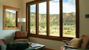 Ein Zimmer mit einem großen Fenster und Blick auf eine gebirgige Landschaft. Es gibt einen Stuhl, einen Schreibtisch mit einem Laptop und eine Lampe.