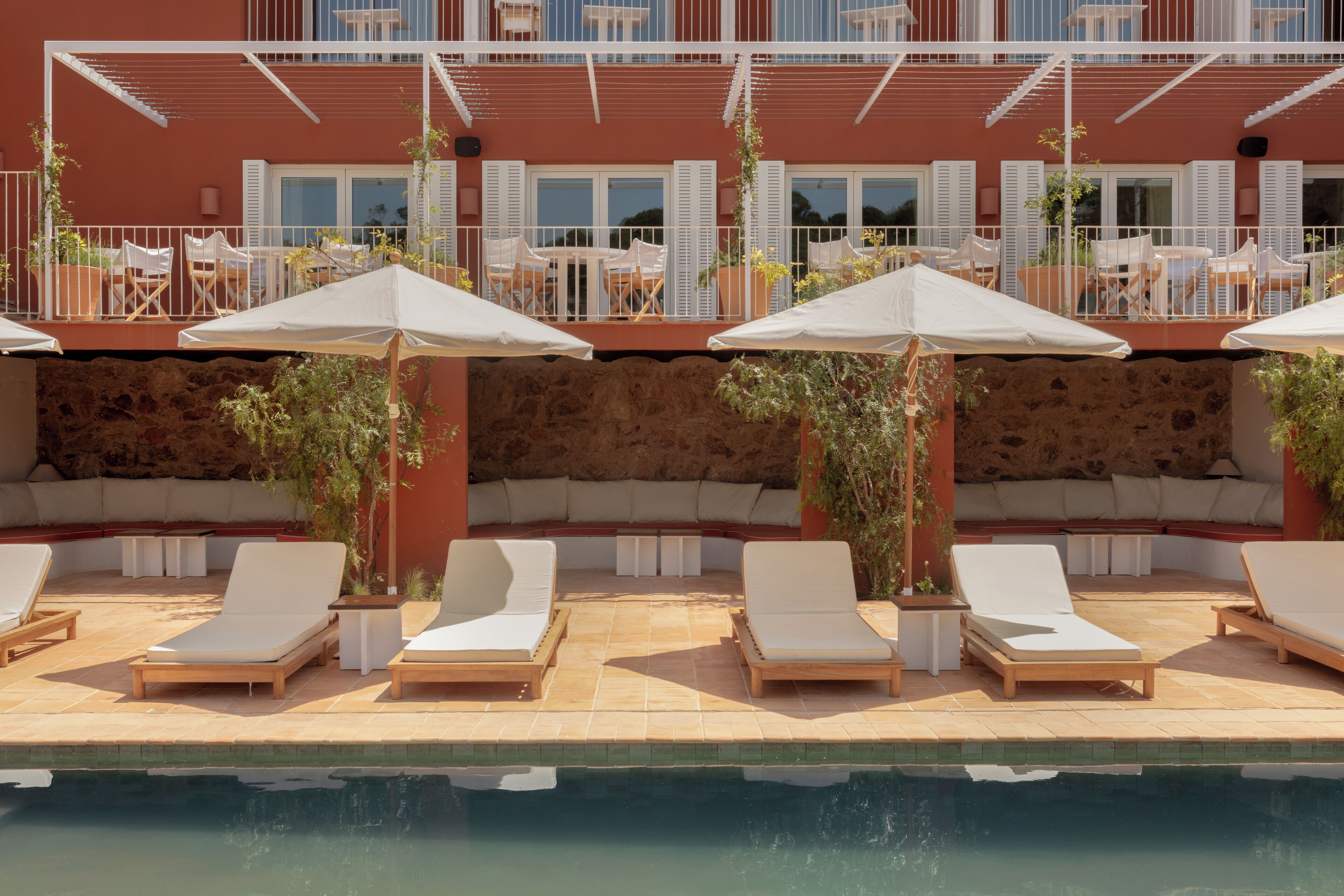A poolside area with lounge chairs and umbrellas, a stone wall, and a building with balconies and windows in the background.