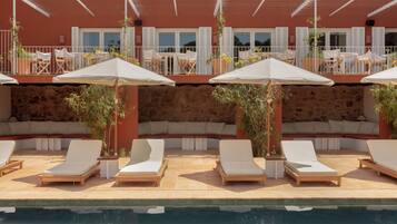 A poolside area with lounge chairs and umbrellas, a stone wall, and a building with balconies and windows in the background.